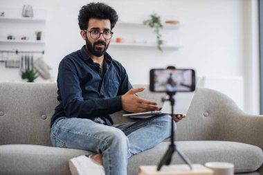 Ciddi bir Arap adam, günlük giysiler içinde gri koltukta oturuyor ve modern akıllı telefondan video kaydederken kablosuz bilgisayarla iletişim kuruyor. Halk ve sosyal medya kavramı.