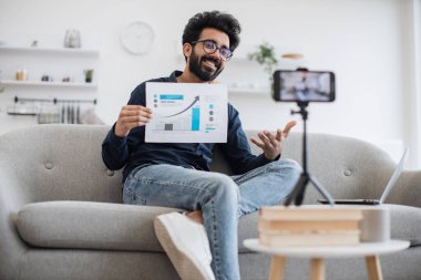 Siyah sakallı genç Arap finansör tripoda sabitlenen cep telefonuyla kendini filme çekiyor ve oturma odasındaki abonelere finansal istatistikler gösteriyor. Teknoloji ve blog kavramı.