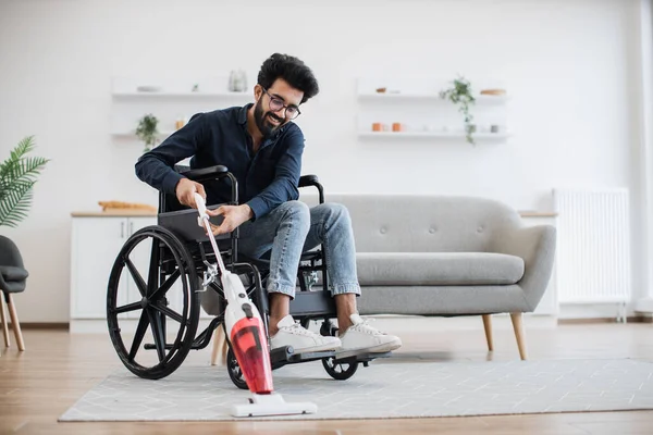 Relaxed adult person in jeans taking lightweight stick vacuum while using wheelchair in spacious flat. Cheerful indian homeowner enjoying housework with modern appliances while waiting for guests.