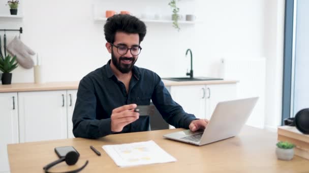 キッチンのインテリアにポータブルコンピュータを入力しながら 空白のクレジットカードを保持眼鏡で魅力的な髭の男 リモートで作業しながらオンライン銀行取引を行う幸せなアラビア人紳士 — ストック動画