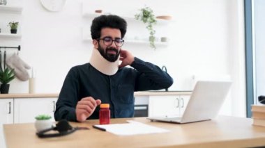 Hintli bir yetişkinin boynu için ağrı kesici tutarken parlak iş yerindeki yazı masasında boyun tasmasını düzeltiyorum. Genç girişimci kaza nedeniyle şirket ofislerini ev bazlı işe çevirdi..