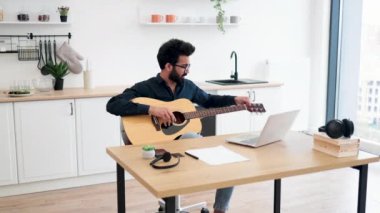 Koyu renk gömlekli, akustik gitar akort eden genç bir müzisyen modern dairede masa başında dizüstü bilgisayar kullanıyor. Çevrimiçi öğretim yoluyla telli enstrüman üzerinde ses egzersizleri yapan odaklanmış Hintli erkek..