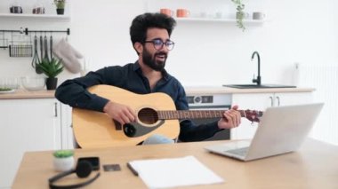 Geniş apartmandaki taşınabilir bilgisayar webcam 'ine gülümserken telli çalgılar tutan neşeli bir Arap. Mutlu gözlüklü erkek modern teknolojileri kullanarak online gitar dersi veriyor..