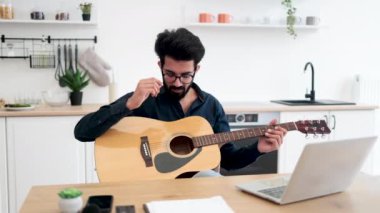 Yetenekli Hintli müzisyen elinde akustik gitar tutarken yemek odasındaki dizüstü bilgisayardaki bilgileri analiz ediyor. Genç sakallı besteci dijital cihazlar ve enstrümanlar kullanarak yeni şarkı yazıyor.