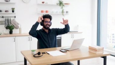 Mutfak masasında dijital aletlerle otururken elleri havada melodiden melodiye hareket ettiren heyecanlı genç adam. Tutkulu Hintli yetişkin kablosuz kulaklıklarla sunulan zengin ses deneyimi.