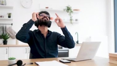 Mutfak masasında dijital aletlerle otururken elleri havada melodiden melodiye hareket ettiren heyecanlı genç adam. Tutkulu Hintli yetişkin kablosuz kulaklıklarla sunulan zengin ses deneyimi.