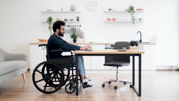 在现代公寓开放式厨房的写字台上工作的坐在轮椅上的印度男性侧视图画像 残疾青年从工作日开始在家中检查电子邮件帐户 — 图库视频影像