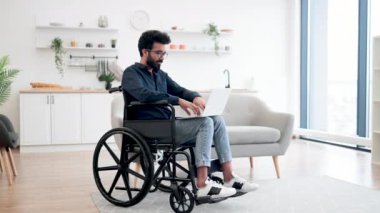Tekerlekli sandalyedeki neşeli Hintli adam dizüstü bilgisayar tutarken açık-plan mutfağın ortasında kalıyor. Evde geçirdiği kazadan sonra iyileşme sürecinde internet sayfalarında mutlu genç sörfçüler..