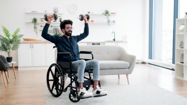 車椅子でハンサムなインド人男性の完全な長距離ビューは室内で限られた可動性を行使しながら重みを持ち上げる 台所の家の試しの間の適度な強度の活動を使用して笑顔の大人 — ストック動画