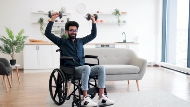 車椅子でハンサムなインド人男性の完全な長距離ビューは室内で限られた可動性を行使しながら重みを持ち上げる 台所の家の試しの間の適度な強度の活動を使用して笑顔の大人 — ストック動画