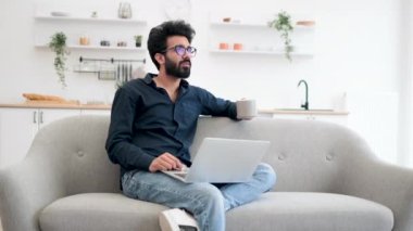 Neşeli Hintli olgun insan portatif bilgisayar ekranında gülümserken açık planlı mutfakta sıcak kahve içiyor. Gözlüklü şık adam sosyal medya sitesinde otururken evde eğleniyor..