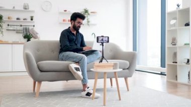 Modern akıllı telefondaki takipçilere günlük kıyafet kayıt videosundaki neşeli içerik yapıcının ön görüntüsü. Dizüstü bilgisayarlı genç Hintli adam oturma odasında rahat bir koltukta oturuyor..