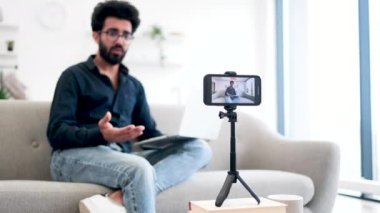 Kanepede dizüstü bilgisayarla oturan ve akıllı telefon kamerasıyla konuşan neşeli bir Arap 'ın bulanık arka planı. Sosyal medyası için olumlu erkek blogcu video kaydı.