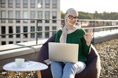 Portatif bilgisayar ve terasta rahat bir armut çantayla poz veren tesettürlü güzel bir Arap kadın. İyi organize olmuş genç bir çalışan açık havanın keyfini çıkarırken gün ortası tabelasında boş duruyor.