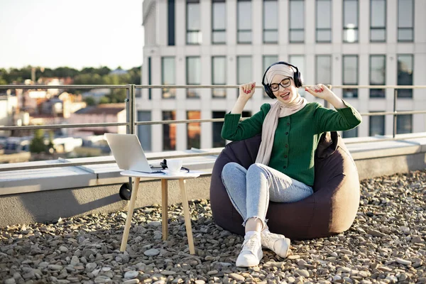 Stock image Easygoing lady in traditional muslim scarf moving to beat of songs in headphones on panoramic terrace with laptop. True music lover relishing pure performance delivered by wireless device outdoors.