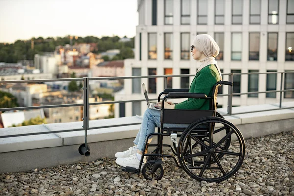 stock image Side view of muslim lady in headscarf using digital device for remote work on panoramic terrace. Well -organized businesswoman in wheelchair searching information over internet application outdoors.