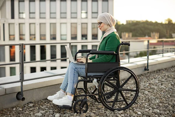 Türbanlı ve gözlüklü müslüman bir kadın modern aleti terasta dizlerinin üzerinde tutarken dizinde daktilo kullanıyor. Tekerlekli sandalyedeki verimli serbest çalışan dışarıda sanal bir iş yapıyor..