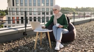 Portatif bilgisayarlarda web sayfalarını kaydırırken başörtüsü içinde elinde bir bardak sıcak içecek tutan çekici Müslüman kadın. Çatıda kahve molası sırasında online alışveriş yapmaktan zevk alan mutlu bir kadın..