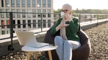 Tesettürlü genç bir kadının portresi kahve içerken terastaki puf minderinin yanına dizüstü bilgisayar koyuyor. Gündelik giysiler içindeki Arap kadın açık havada profesyonel faaliyetler yürütüyor..