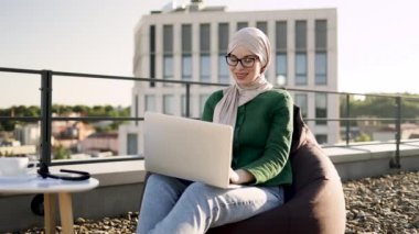 Kahve masasının yanındaki puf minderde oturan tesettürlü, tesettürlü ve açık havada içkili gülümseyen Arap kadın. Gözlüklü neşeli iş kadını işe terasta dizüstü bilgisayarla başlıyor..