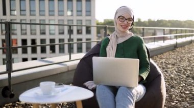 Portatif bilgisayar ve terasta rahat bir armut çantayla poz veren tesettürlü güzel bir Arap kadın. İyi organize olmuş genç bir çalışan gün ortasında açık havanın keyfini çıkarırken,.
