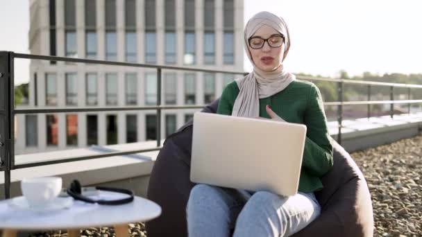 屋上テラスで快適なPouff椅子に座っている間 ポータブルコンピュータを利用してメガネでイスラム教徒の大人を自信を持って 屋外の職場でビデオ通話を取るスカーフの焦点を当てたビジネス女性 — ストック動画