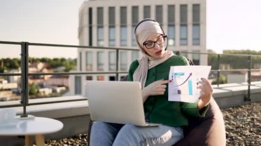 Çevrimiçi toplantı sırasında modern teknolojiler aracılığıyla açık havada yapılan kağıt bilgilerine işaret eden odaklanmış Müslüman yetişkinler. Panoramik terasta finansal veri planını tartışan tesettür içindeki sakin iş kadını.
