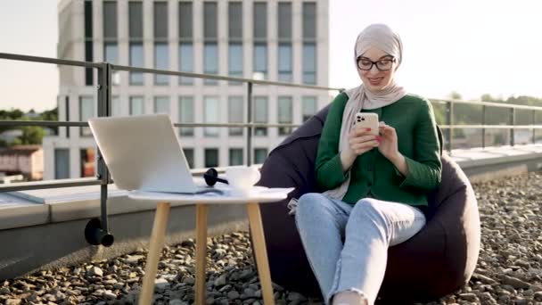 正午にコーヒー休憩中に屋上テラスのPouff椅子に座ってスマートフォンを持つ陽気な人の閉鎖 ヒジャーブの幸せなアラビア語の女性とオープンエアでモバイル経由でアイウェアのテキストメッセージ — ストック動画