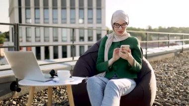 Neşeli Müslüman kadın çatıdaki puf koltukta dinlenirken telefon konuşmasına katılıyor. Tesettürlü çekici bayan kahve içerken iş arkadaşlarıyla sohbet ediyor..