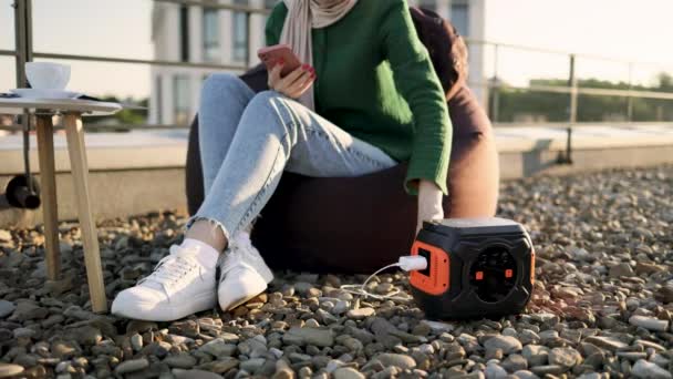 ビーンバッグに座っている間 携帯電話を充電式バッテリーに接続するジーンズの若い女性の作物のビュー 屋外愛好家は オフグリッド電気のおかげでスマートフォンの使用の利点を楽しんで — ストック動画