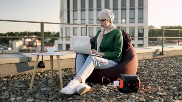 迷人的穆斯林女士戴着头巾 一边摸着手提电脑充电器 一边看着屋顶上的数字屏幕 轻松的女人上网看电影 同时还能上网上网和上网 — 图库视频影像