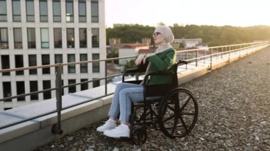 Geleneksel türbanlı çekici Müslüman kadın sabah saatlerini panoramik terasta geçiriyor. Düz çatıdaki kentsel manzaraya tepeden bakan günlük giysiler ve gözlük giyen rahat tekerlekli sandalye kullanıcısı..