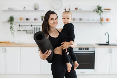 Elinde yoga minderiyle neşeli beyaz kadın portresi. Geniş bir mutfakta duran küçük kızı taşıyor. Mutlu anne ve güzel kız atletizmde giysi planlama evinde çalışıyor..
