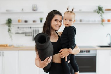 Elinde yoga minderiyle neşeli beyaz kadın portresi. Geniş bir mutfakta duran küçük kızı taşıyor. Mutlu anne ve güzel kız atletizmde giysi planlama evinde çalışıyor..