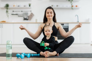 Yoga pozisyonunda oturan rahat bir kadının modern apartmandaki lastik minder üzerinde küçük bir çocukla tam boy görüntüsü. Güzel bir kız dambılları denerken anne, Gyan Mudra 'da parmaklarıyla egzersiz yapıyor..