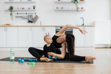 Elinde dambıl olan küçük bir çocuğa bakarken yoga minderinde kollarını ve bacaklarını esneten zayıf esmer kadın. Spor giyim sektöründe genç bir ebeveyn ve kız antrenman sırasında sözlü olmayan iletişimi sürdürüyor..