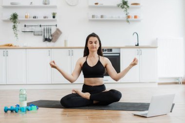 Siyah aktif giysili rahat bir kadın Lotus 'ta meditasyon yaparken elleri Gyan Mudra' da dizüstü bilgisayardan müzik eşliğinde. Çekici bayan, modern apartmanda yoga yaparken dinlenme melodilerini seviyor..