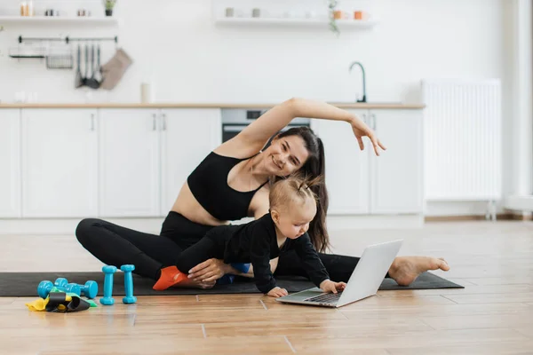 stock image Smiling caucasian woman in active wear exercising while little daughter touching keyboard of laptop on floor. Happy female parent boosting fitness while entertaining baby with technologies at home.