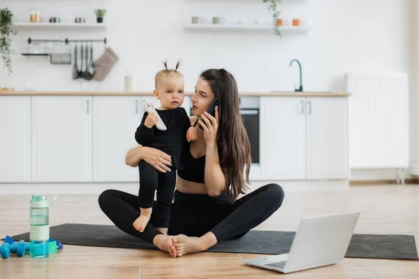 stock image Charming woman with cell phone sitting cross-legged on yoga mat with little child on hip in kitchen interior. Busy mother speaking on mobile during exercise while cute daughter holding gadget in hand.