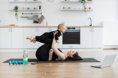 Atletik ince kadın, mutfak içinde lastik paspasın üzerinde yatarken dizlerinin üzerinde bebek kızı taşıyor. Spor elbiseli kadın ebeveyn evde kızıyla eğlenirken dayanıklılığını arttırıyor..