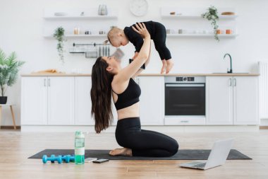 Aktif giyimde neşeli bir ebeveyn, yoga minderinin altında bacakları katlanmış bir şekilde otururken havada küçük bir çocuk yetiştiriyor. Neşeli narin anne egzersizleri evde oyuna çevirirken dengesini geliştiriyor..