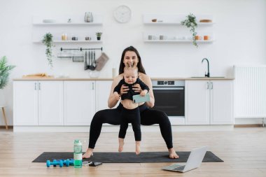 Atletik giyimli beyaz tenli çekici bir yetişkin mutfak içinde bebek ellerindeyken geniş çömelme hareketleri yapıyor. Aktif genç anne, güneşli bir günde küçük tatlı kızını kapalı mekanlarda fiziksel aktivitelere sokuyor..