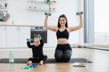 Gülümseyen anne ve bebeğin spor kıyafetleriyle mavi dambılları oturdukları yerden kaldırışlarının tam boy görüntüsü. Neşeli aile kadını evde jimnastik yaparken çocukların gücünü arttırıyor..