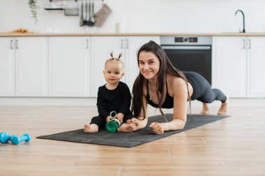 Spor şişesiyle gülümseyen küçük bir kızın yanında kauçuk hasırla poz veren neşeli beyaz kadın. Tarz sahibi bir anne, evde bedeni güçlendirirken kızıyla güçlü bir ilişki kurar..