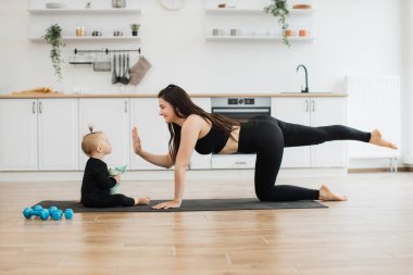 Güzel bir kadının evde yoga yaparken güzel bir kıza beşlik çakması. Spor kıyafetleri içinde atletik bir anne her gün antrenmanı mutfak arka planında oynamaya çeviriyor..