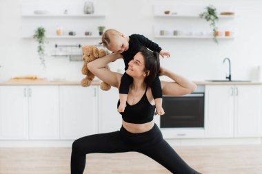 Çekici, zayıf bir kadın evde yoga pozisyonunda gerinirken omuzlarında tutulan çocukla görsel temas kuruyor. Sağlıklı anne, kızını ve yumuşak ayısını taşıyarak fazladan ağırlık kazanıyor..