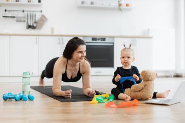 Tatlı çocuk oyuncak ayı için direnç bandı rengini seçerken atletik kadın minder üzerinde tahta poz veriyor. Yoga kıyafetli sportif anne-kız iç mekanlarda kaynaşma deneyimi kazanıyorlar..