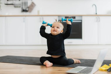 Siyah spor giyim giymiş heyecanlı bir kız elinde halterlerle mutfak arka planındaki lastik paspasın üzerinde dinleniyor. Oda zeminindeki dizüstü bilgisayarın yanında ağırlık kaldırma egzersizlerini taklit eden oynak bir bebek..