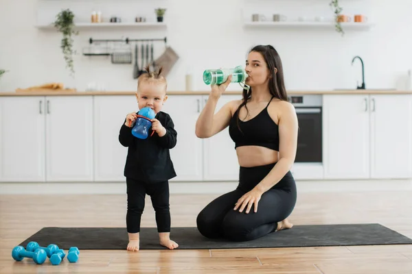 stock image Beautiful adult mom in sports clothes having sip of water while sitting on knees on yoga mat in room. Barefoot little girl posing with baby bottle near sporty mom having break in training at home.