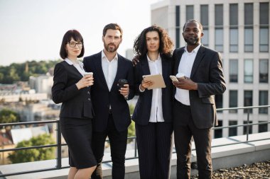 Dışarıda kahve fincanları ve modern aletlerle kameraya poz veren dört uzmanın portresi. Birliktelik ve başarı gösteren çok ırklı çalışanlar. Takım çalışması ve işbirliği kavramı.